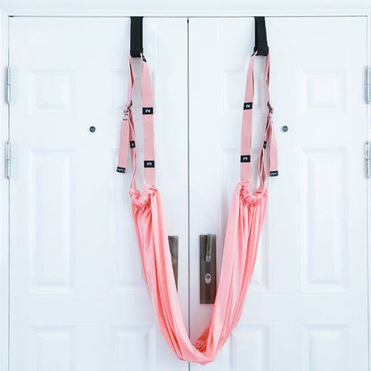 Pink fitness hip stretch yoga belt hanging on double white doors with adjustable straps and foam handles for inverted stretching exercises